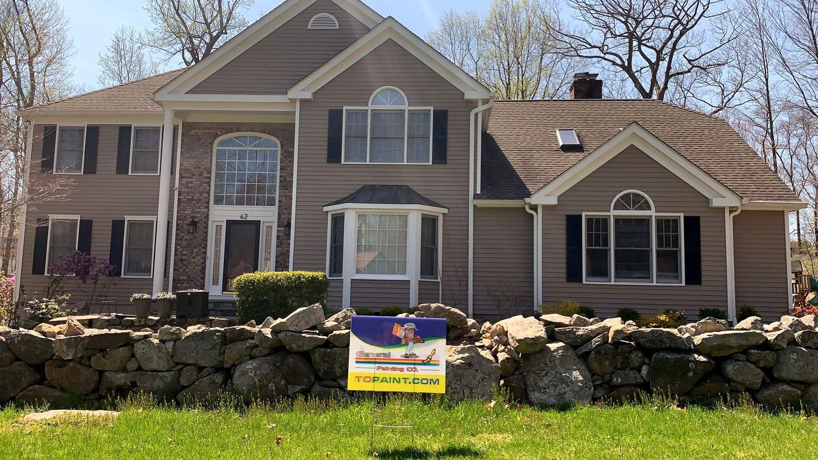 Beige colonial home with stone wall and yard sign