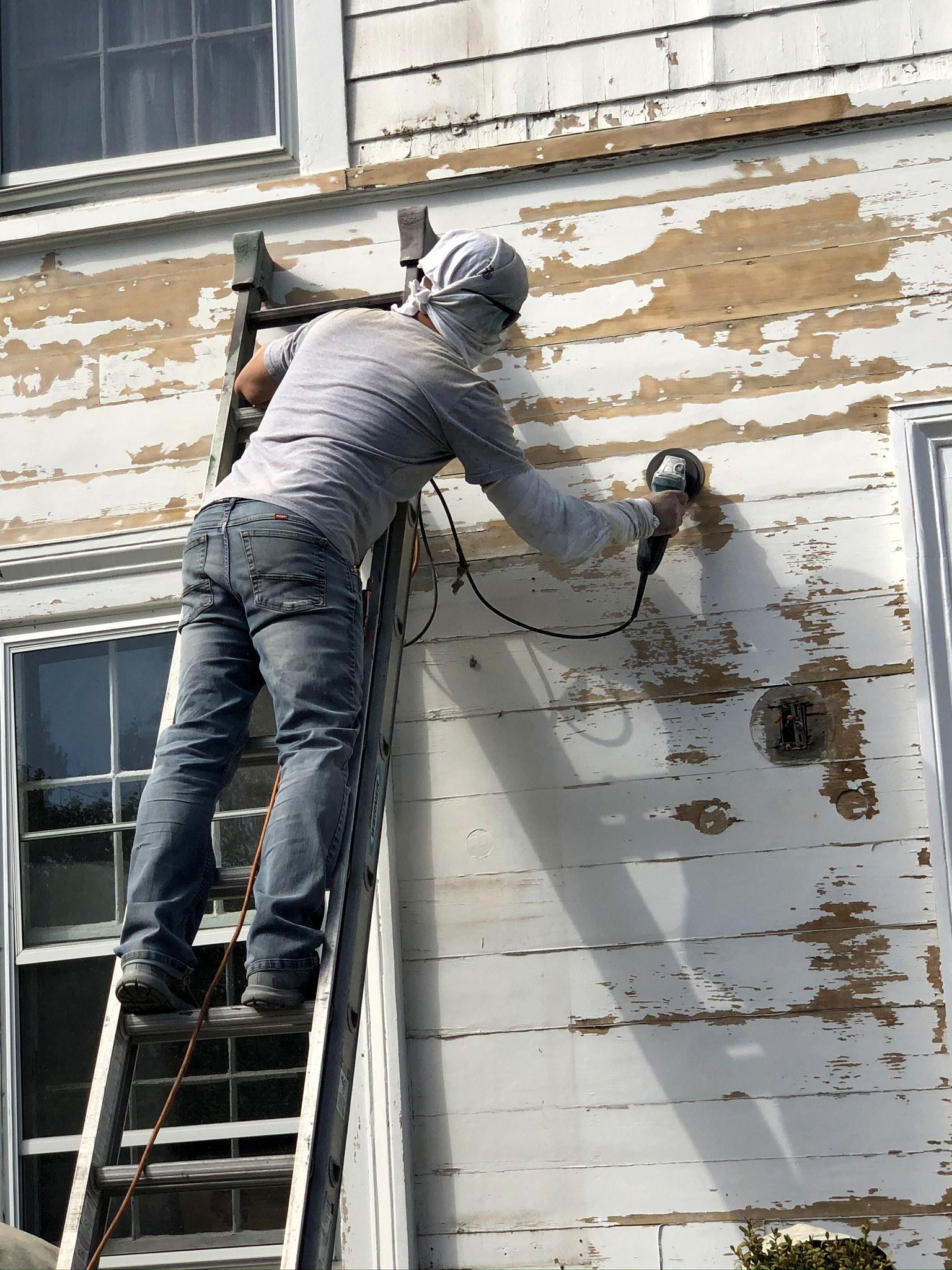 Worker hand-scraping failed paint from white siding — prep stage